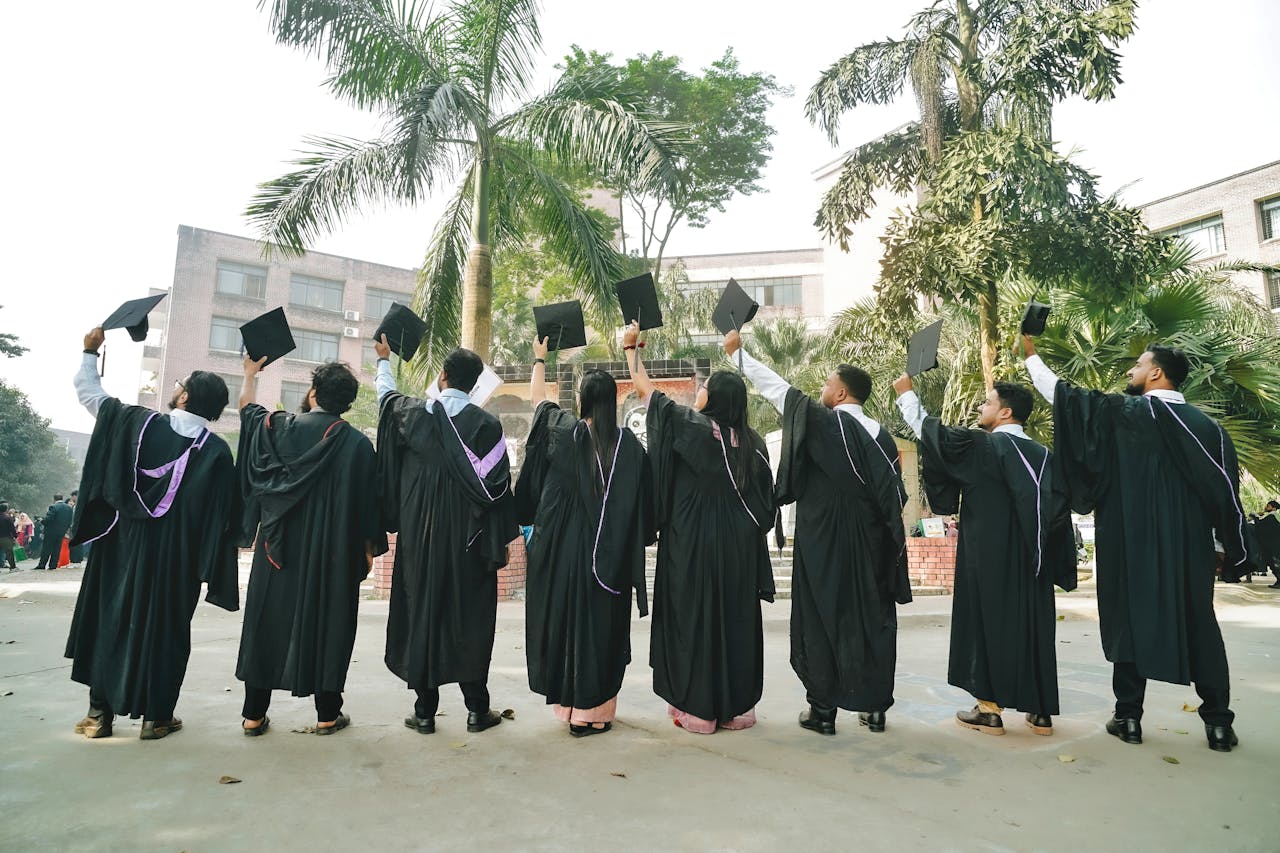A group of graduates wearing gowns celebrate by holding caps high in an outdoor ceremony.