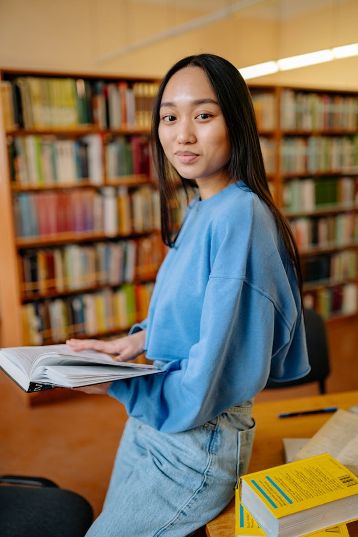 services-02 Asian woman reading a book in a library, smiling while studying surrounded by shelves.
